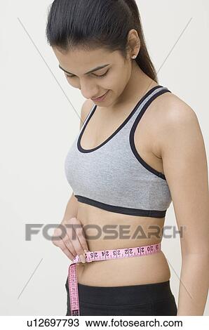 Stock Image - Side profile of a young woman measuring her waist with a tape measure. Fotosearch