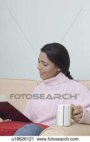Stock Image - Side profile of a young woman sitting on a couch and reading a book. Fotosearch