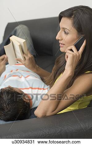 Stock Image - Side profile of a young woman talking on a mobile phone with a young man reading a book. Fotosearch
