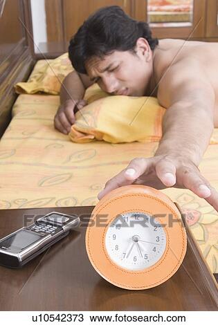 Young man lying on a bed and reaching for an alarm clock View Large Photo Image Stock Image - Young man lying on a bed and reaching for an alarm clock. Fotosearch