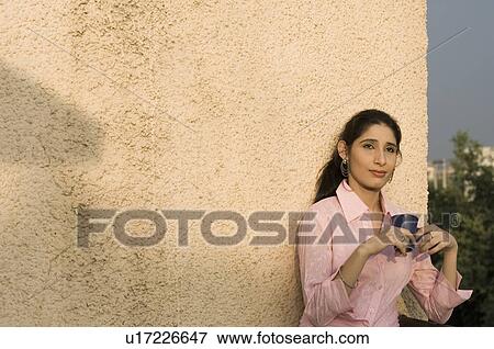Stock Photo - Young woman leaning against a wall and holding a cup of coffee. Fotosearch