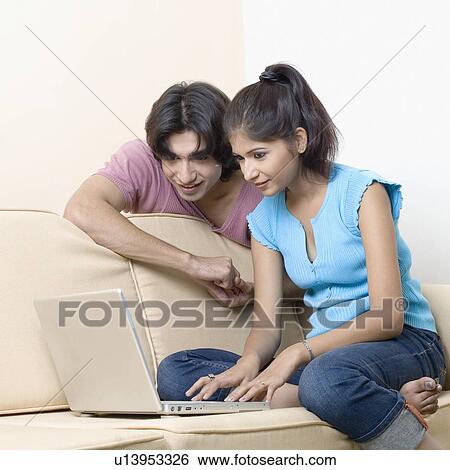 Young woman sitting on a couch and using a laptop with a young man beside her View Large Photo Image Stock Photograph - Young woman sitting on a couch and using a laptop with a young man beside her. Fotosearch