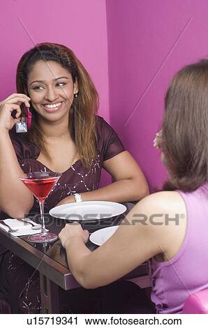 Young woman sitting with her friend in a restaurant and using a mobile phone View Large Photo Image Stock Image - Young woman sitting with her friend in a restaurant and using a mobile phone. Fotosearch