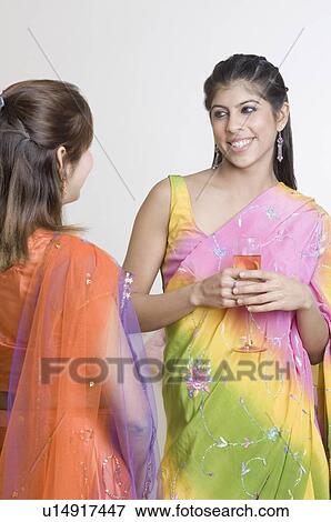 Stock Photo - Young woman standing holding a glass of drink and looking at another young woman. Fotosearch
