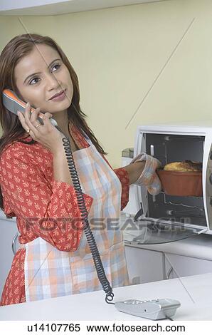 Stock Photography - Young woman talking on the telephone and taking a cake out of the oven. Fotosearch