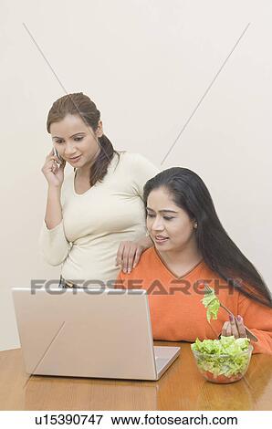 Stock Photo - Young woman using a laptop with another woman beside her talking on a mobile phone. Fotosearch