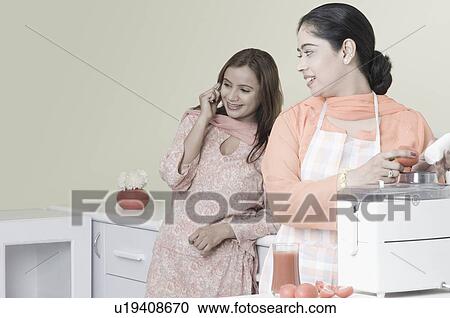 Stock Image - Young woman using an electric mixer with another woman talking on a mobile phone. Fotosearch