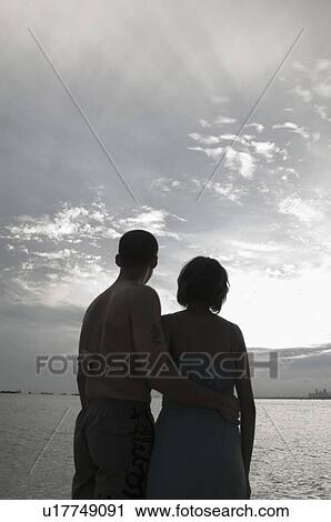 Stock Image - Rear view of a couple standing on the beach with their arms around each other. Fotosearch