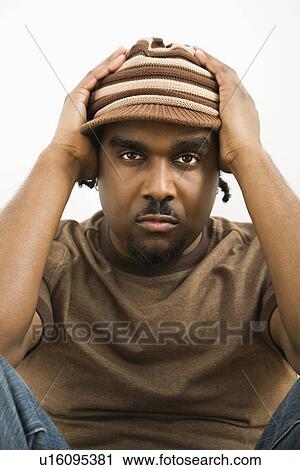 Stock Image - African-American mid-adult man holding head wearing hat looking at viewer.. Fotosearch
