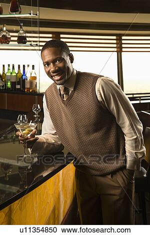 Stock Image - African American mid adult man leaning on bar with martini looking at viewer.. Fotosearch