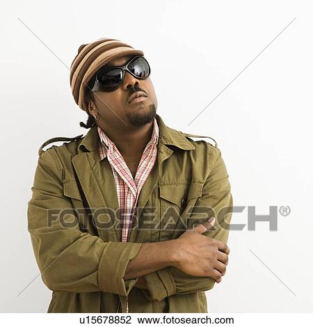 Stock Image - African-American mid-adult man wearing hat and sunglasses with arms crossed looking up.. Fotosearch