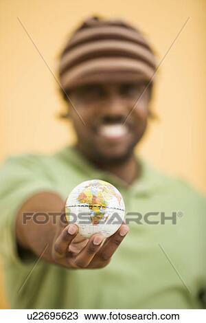Stock Image - African-American mid-adult man wearing hat holding small globe to viewer.. Fotosearch