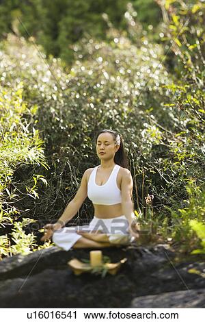 Stock Image - Asian American woman sitting on boulder in forest meditating with candle in Maui, Hawaii.. Fotosearch