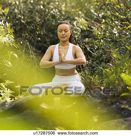 Stock Photograph - Asian American woman sitting on boulder in forest meditating with candle in Maui, Hawaii.. Fotosearch