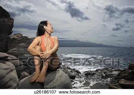 Stock Photo - Asian woman sitting on rock by ocean looking over shoulder smiling in Maui, Hawaii. Fotosearch