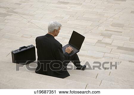 Back view of Caucasian middle aged businessman typing on laptop with briefcase sitting on steps outdoors. View Large Photo Image Stock Image - Back view of Caucasian middle aged businessman typing on laptop with briefcase sitting on steps outdoors.. Fotosearch