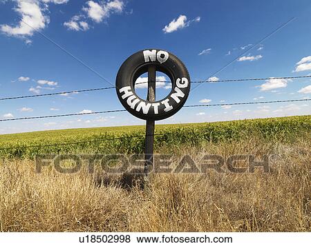 Stock Photo - Barbed wire fence with tire reading No Hunting in front of agricultural field.. Fotosearch