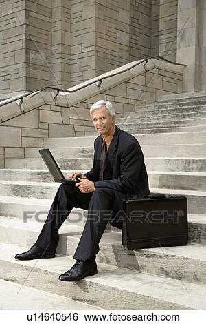 Stock Photograph - Businessman sitting on steps outdoors with laptop and briefcase. Fotosearch