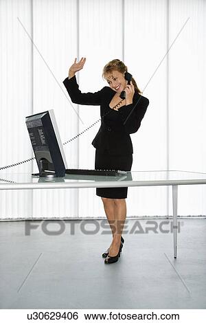 Businesswoman standing at computer desk waving on telephone View Large Photo Image Stock Photograph - Businesswoman standing at computer desk waving on telephone. Fotosearch