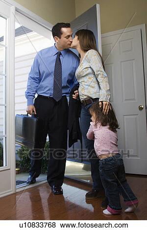 Stock Photo - Caucasian businessman at open door kissing wife while daughter hugs her leg.. Fotosearch