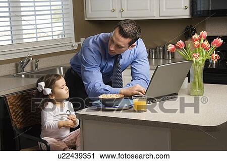 Stock Image - Caucasian father in suit using laptop computer with daughter eating breakfast in kitchen.. Fotosearch