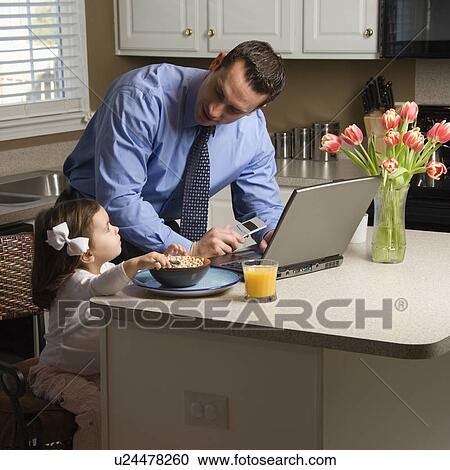 Stock Image - Caucasian father in suit using laptop computer with daughter eating breakfast in kitchen.. Fotosearch