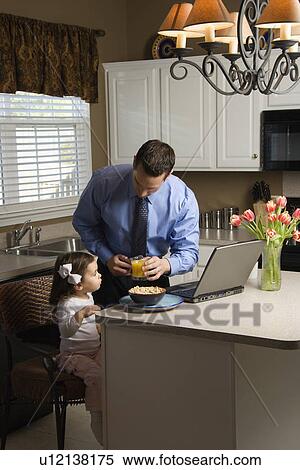 Stock Photography - Caucasian father in suit using laptop computer with daughter eating breakfast in kitchen.. Fotosearch