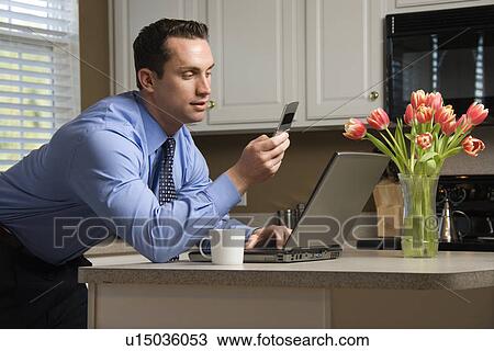 Stock Image - Caucasian man in suit using laptop computer and cellphone in kitchen.. Fotosearch