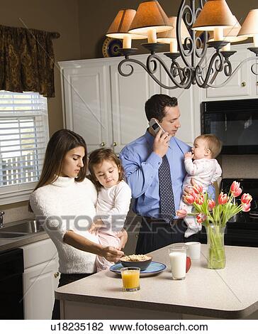 Stock Image - Caucasian mother and father in kitchen busy with children and cellphone.. Fotosearch