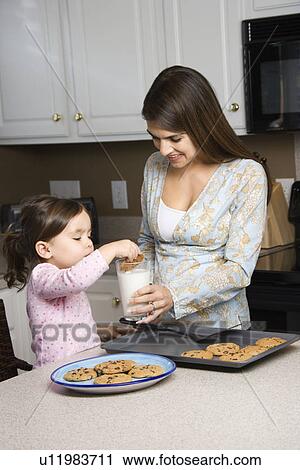 Stock Image - Caucasian mother holding milk glass while daughter dunks cookie.. Fotosearch