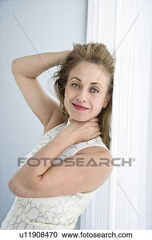 Stock Image - Caucasian woman leaning on wall smiling at viewer with hands touching head.. Fotosearch