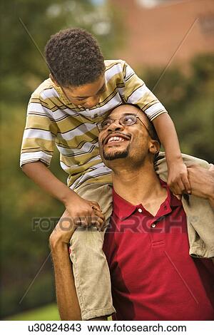 Stock Image - Father carrying his son on his shoulders smiling and looking at eachother.. Fotosearch