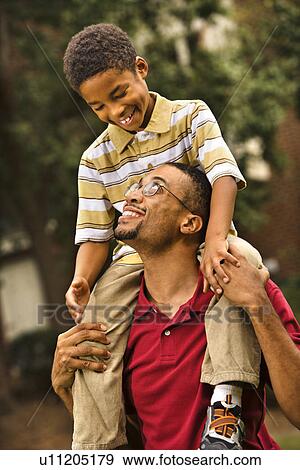 Stock Photo - Father carrying his son on his shoulders smiling and looking at each other. Fotosearch