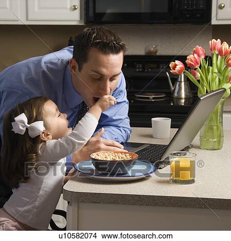 Father in suit using laptop computer with daughter feeding him breakfast in kitchen View Large Photo Image Picture - Father in suit using laptop computer with daughter feeding him breakfast in kitchen. Fotosearch