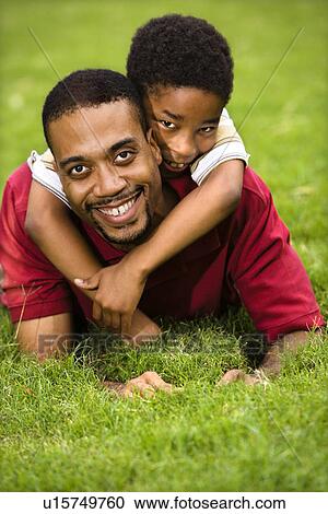 Stock Image - Father laying in grass smiling as son climbs on his back and hugs his neck. Fotosearch