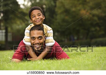 Stock Image - Father lying in grass smiling as son climbs on his back and hugs his neck.. Fotosearch