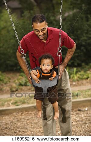 Stock Photograph - Father pushing young son on swing in park in playground. Fotosearch