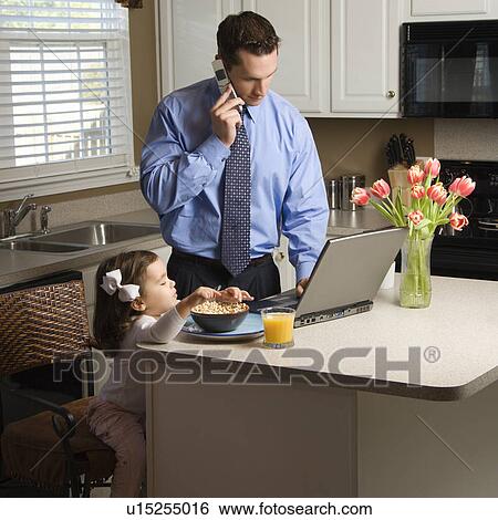 Stock Photograph - Father talking on cellphone and using laptop computer with daughter eating breakfast in kitchen. Fotosearch