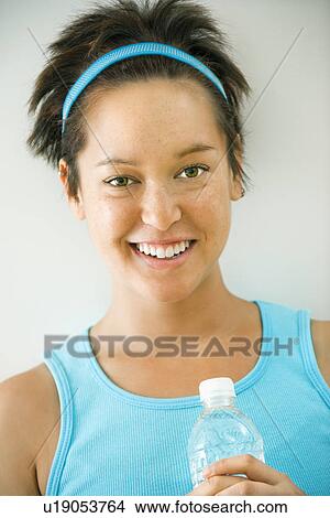 Picture - Head and shoulder portrait of young woman in fitness wear holding bottled water and smiling.. Fotosearch