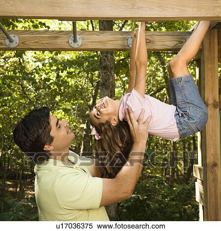 Stock Photography - Hispanic girl hanging by arms and legs from monkey bars smiling at father helping her.. Fotosearch