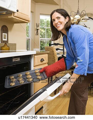 Stock Image - Hispanic mid adult woman putting cookies into oven and smiling at viewer.. Fotosearch