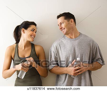 Man and woman at gym in fitness attire holding water bottles standing against wall smiling at eachother. View Large Photo Image Picture - Man and woman at gym in fitness attire holding water bottles standing against wall smiling at eachother.. Fotosearch