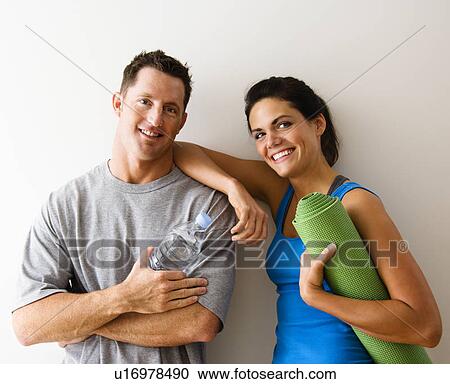 Stock Image - Man and woman at gym in fitness attire holding water bottles and yoga mat standing against wall smiling. Fotosearch