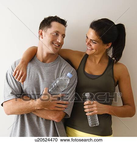 Stock Image - Man and woman at gym in fitness attire holding water bottles standing against wall smiling at eachother.. Fotosearch
