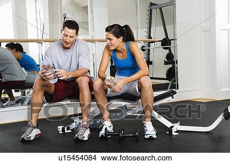 Picture - Man and woman sitting on exercise machine talking holding water bottles. Fotosearch