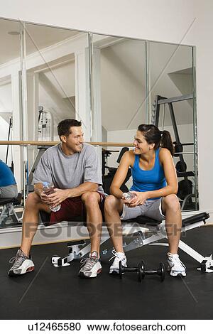 Stock Image - Man and woman sitting on exercise machine talking holding water bottles.. Fotosearch