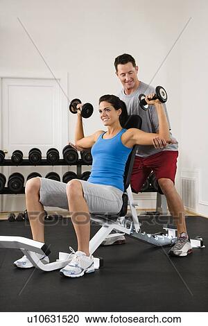 Stock Image - Man assisting woman at gym with hand weights smiling. Fotosearch