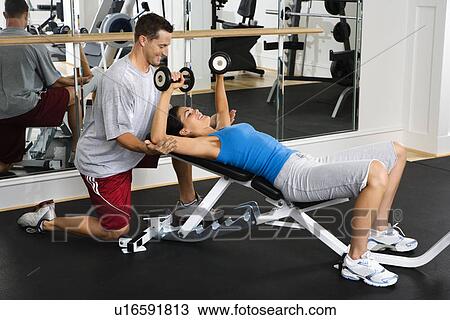 Stock Image - Man assisting woman at gym with hand weights and smiling. Fotosearch
