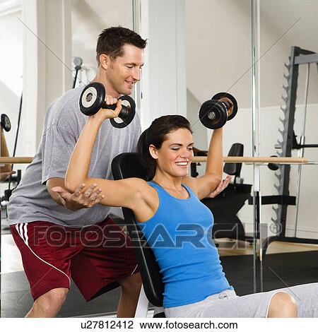 Stock Image - Man assisting woman at gym with hand weights smiling.. Fotosearch