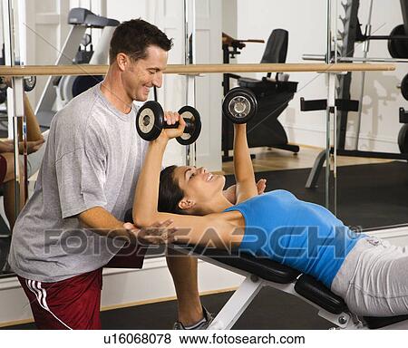 Stock Photo - Man assisting woman at gym with hand weights smiling. Fotosearch